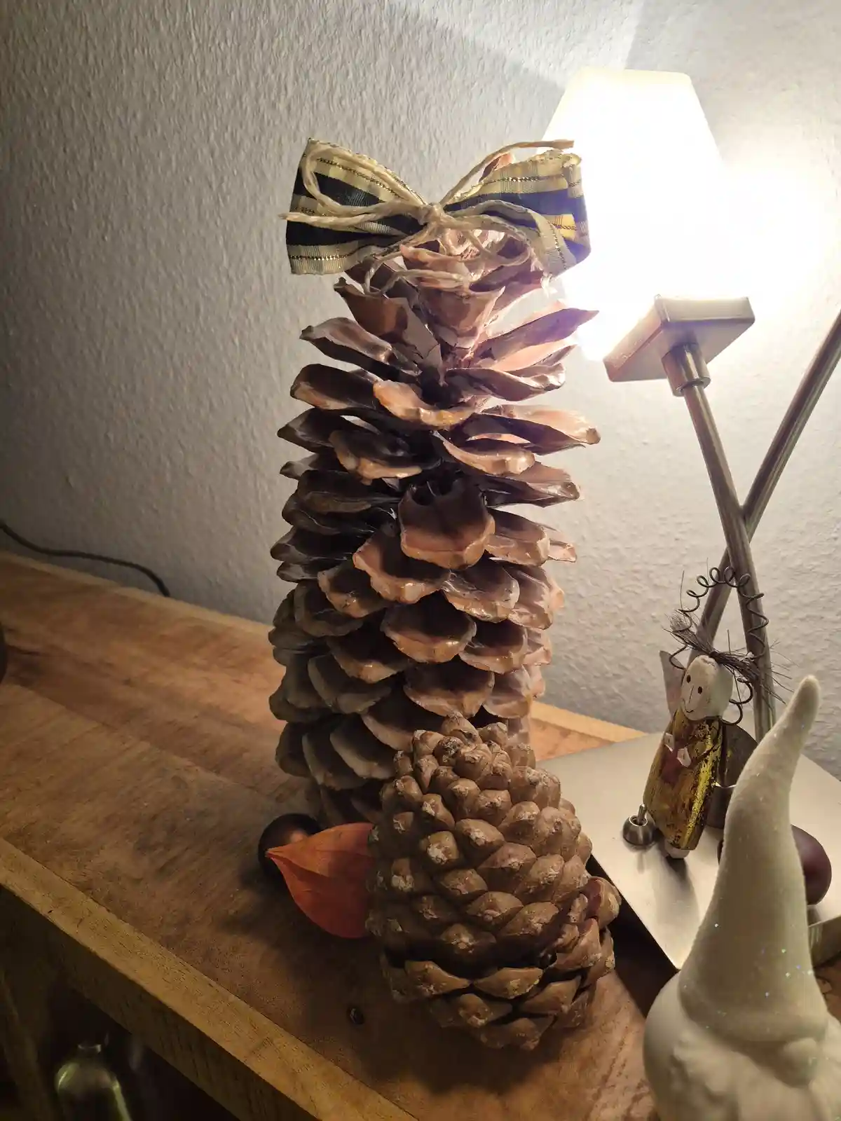 A tall, decorative pine cone tree made of stacked pine cones sits on a wooden table, topped with a plaid ribbon bow, next to…