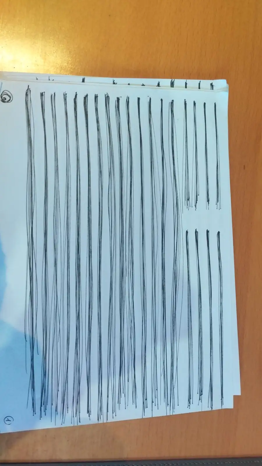 A close-up photo shows a stack of white paper on a wooden surface, covered in numerous messy, uneven vertical black lines…