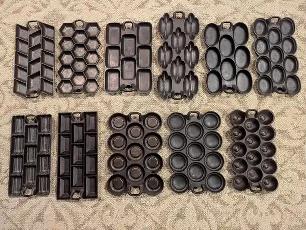 Ten black cast iron cake pans of various shapes and sizes are arranged in two rows on a beige carpet, forming the word…