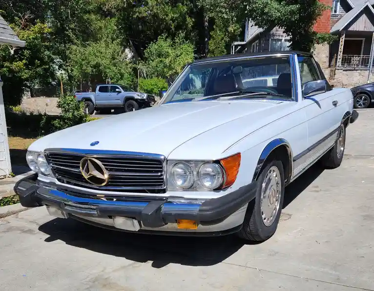 A white 1970s Mercedes-Benz 450SL convertible is parked in a driveway, its chrome grille and round headlights gleaming under…