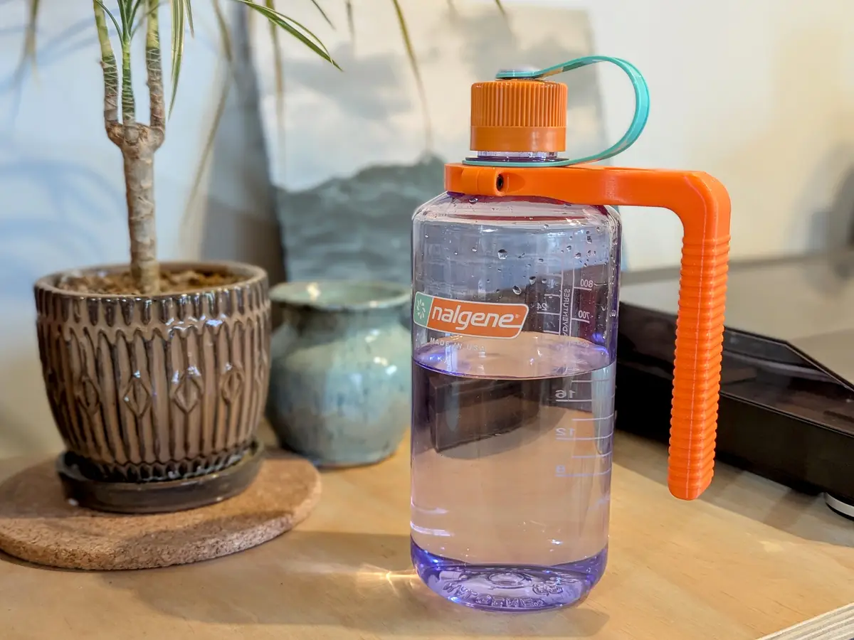 A clear Nalgene water bottle with an orange handle and cap sits on a wooden table, partially filled with water. In the…