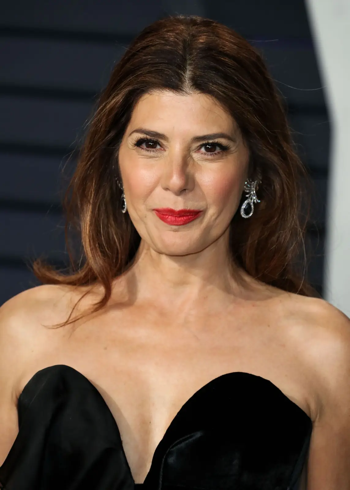 A woman with brown hair and red lipstick wears a black strapless dress and diamond earrings, looking directly at the camera…