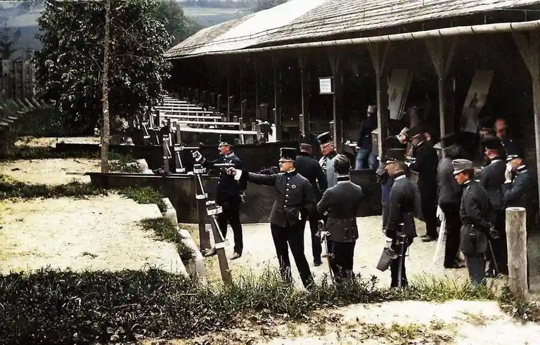 A vintage colorized photograph shows a group of men in early 20th-century military uniforms gathered at an outdoor shooting…