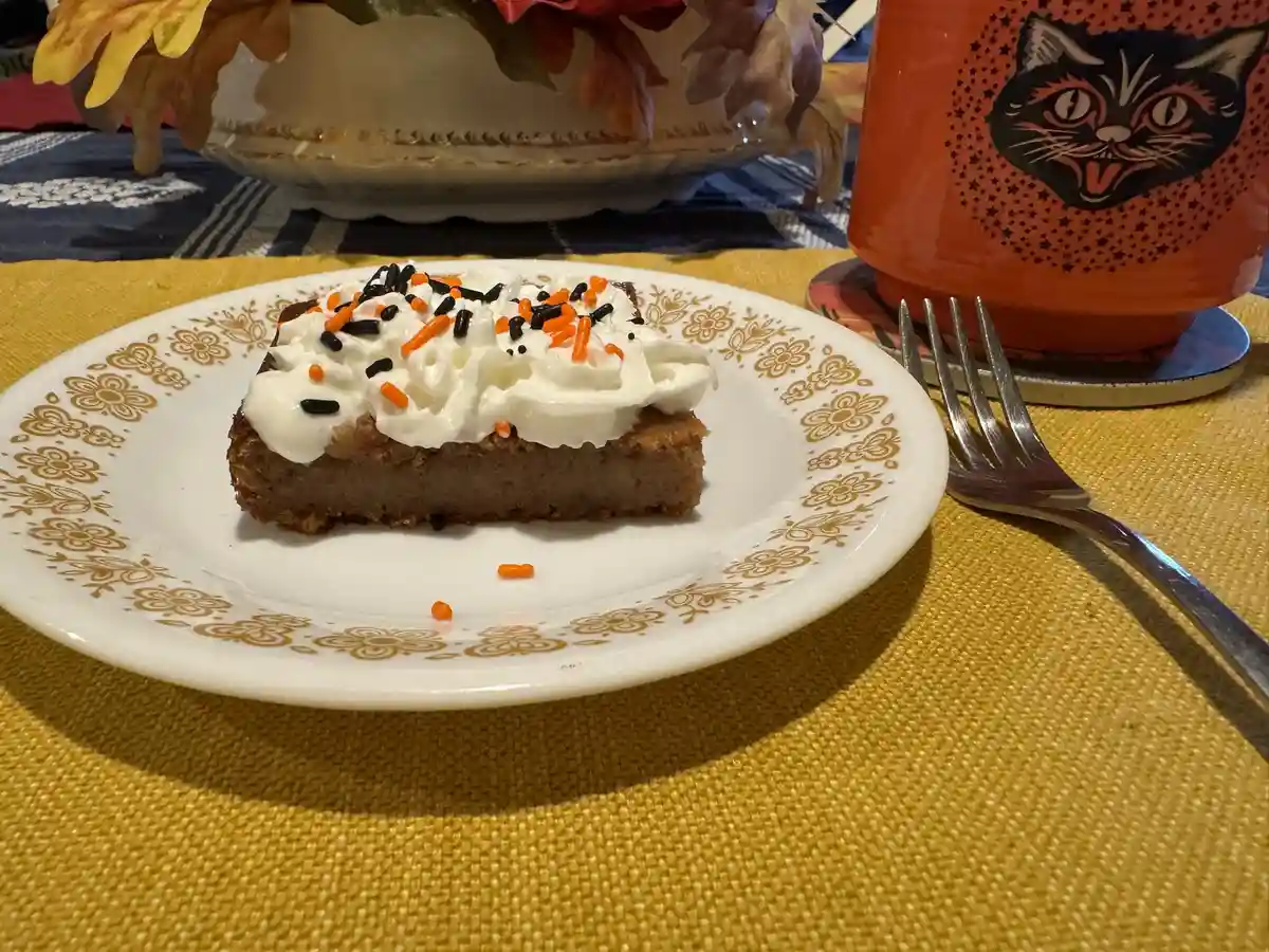 A slice of brown cake with white whipped cream and orange/black sprinkles sits on a white plate with a gold floral rim…