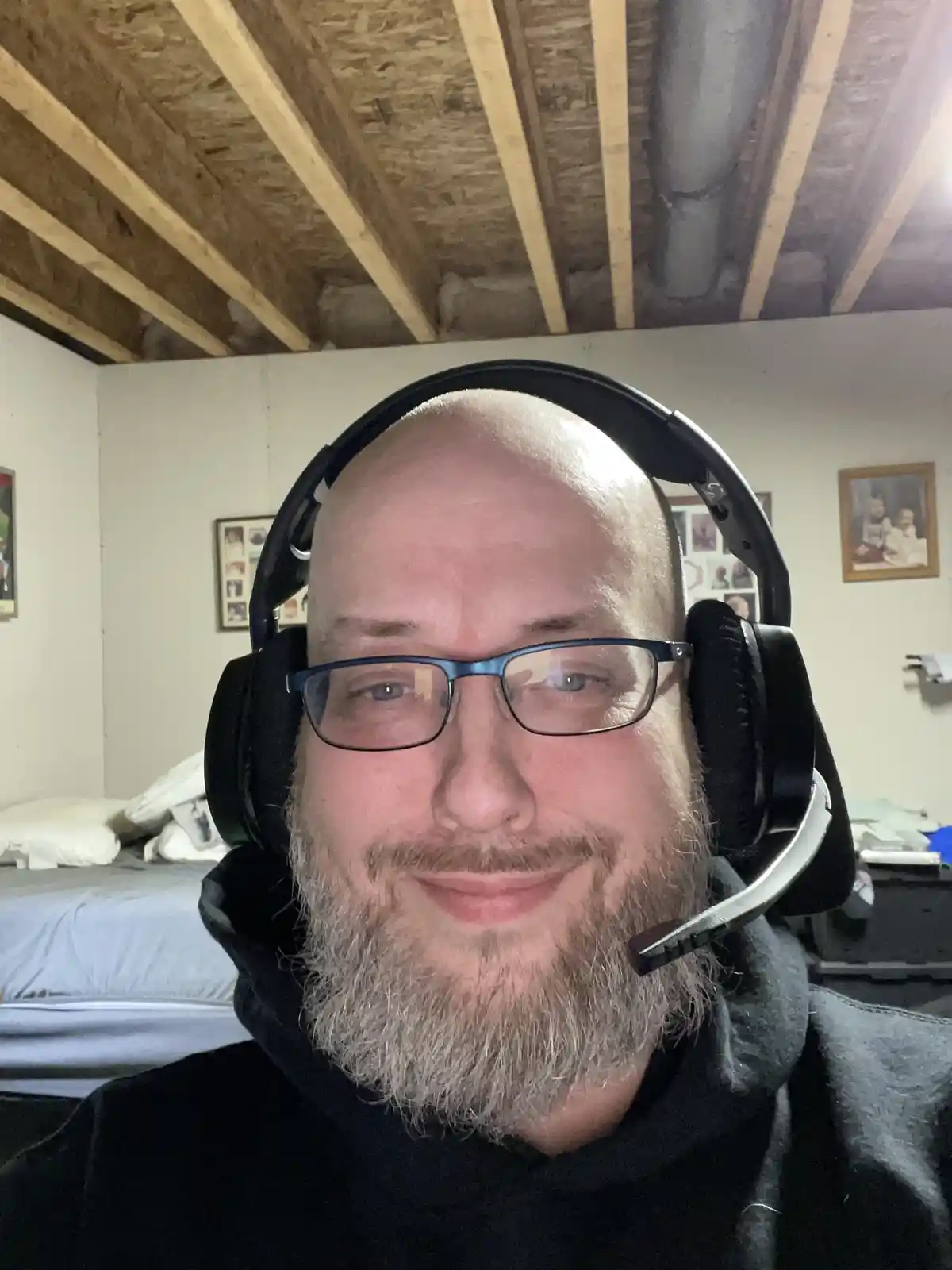 A bald man with glasses and a gray beard wears a black headset with a microphone, smiling slightly at the camera. Behind…