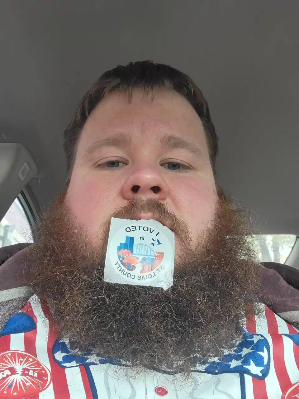 A man with a thick, bushy beard is taking a selfie inside a car, holding a circular 'I VOTED in ST. LOUIS COUNTY' sticker in…