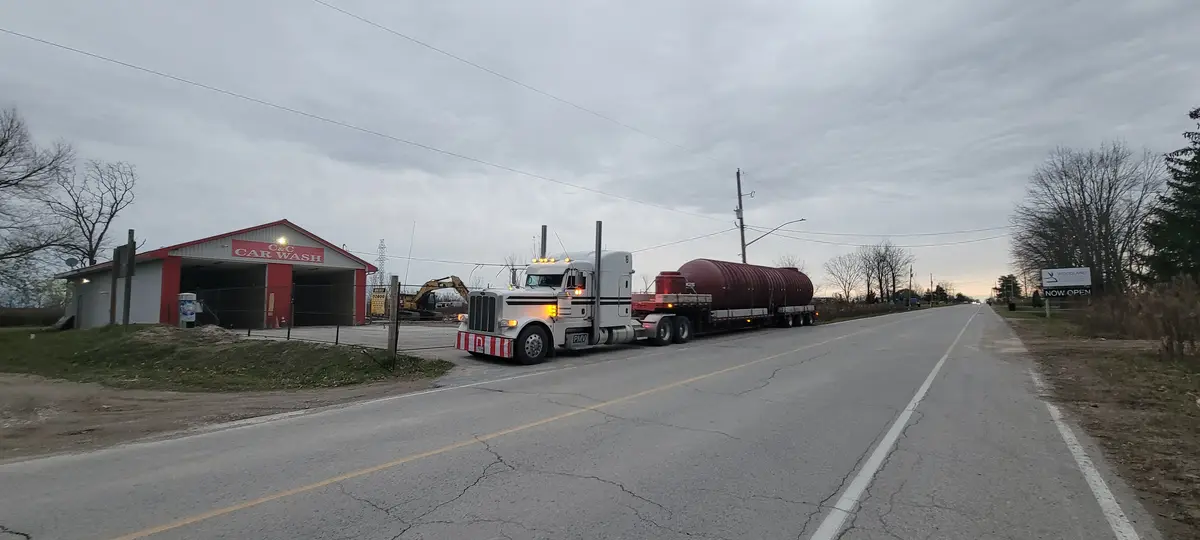 A white semi-truck with a large red tank trailer is stopped on a rural road next to a building labeled 'C&C CAR WASH'. The…