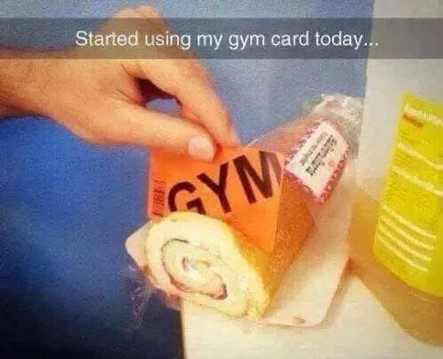 A hand holds an orange card labeled 'GYM' that is stuck into a sandwich wrap, which is wrapped in plastic and sits on a…