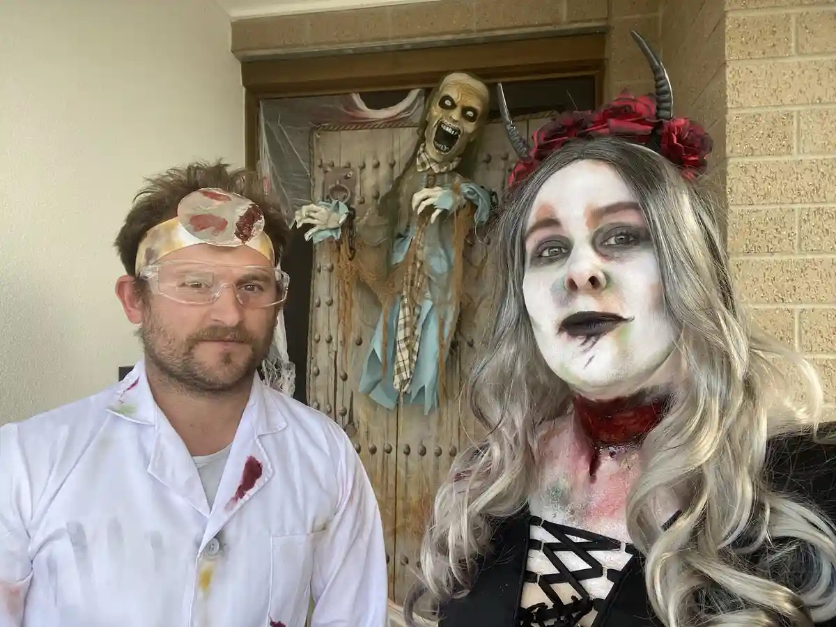 Two people in Halloween costumes pose for a photo: a man in a stained white lab coat with a bloody bandage on his head and…
