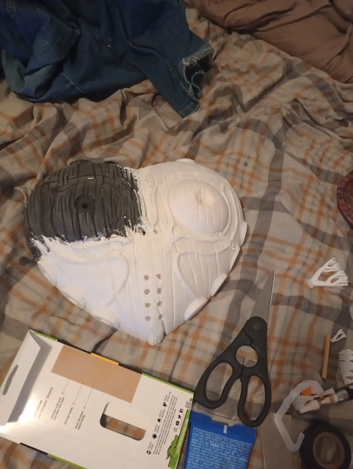 A partially painted white mask, resembling a Star Wars Stormtrooper helmet, lies on a plaid blanket surrounded by crafting…