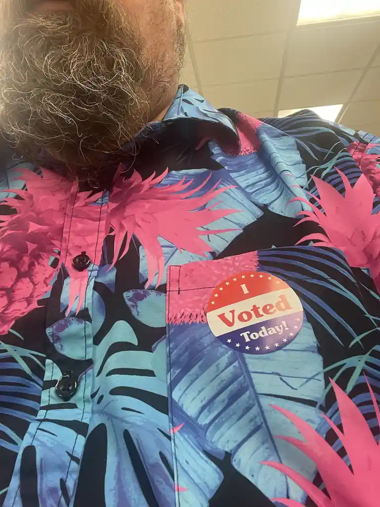 A close-up shot shows the upper torso of a person with a beard wearing a brightly colored Hawaiian shirt with pink and blue…