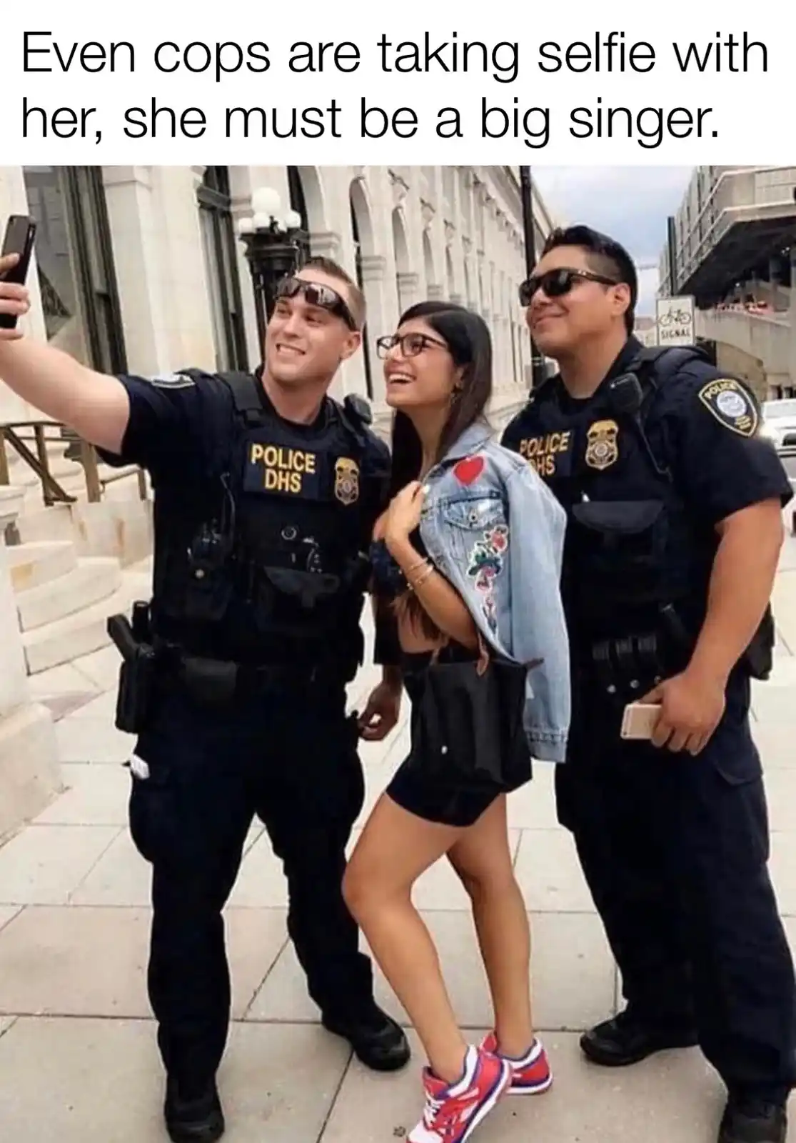 Three people, two police officers and a woman, are posing for a selfie on a city sidewalk. The officer on the left holds up…
