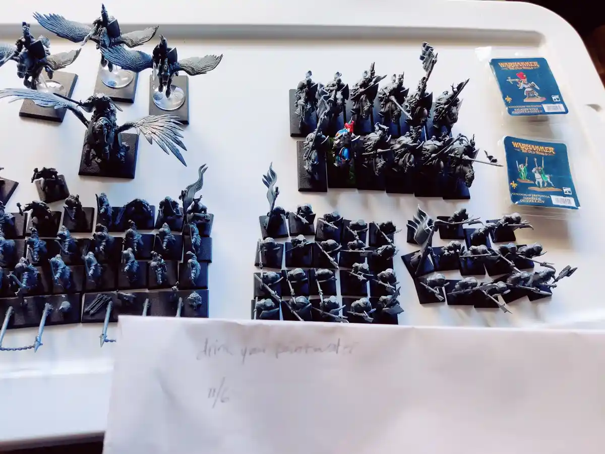 A white tray holds numerous unpainted Warhammer miniatures, including mounted knights and infantry, arranged in rows. Two…