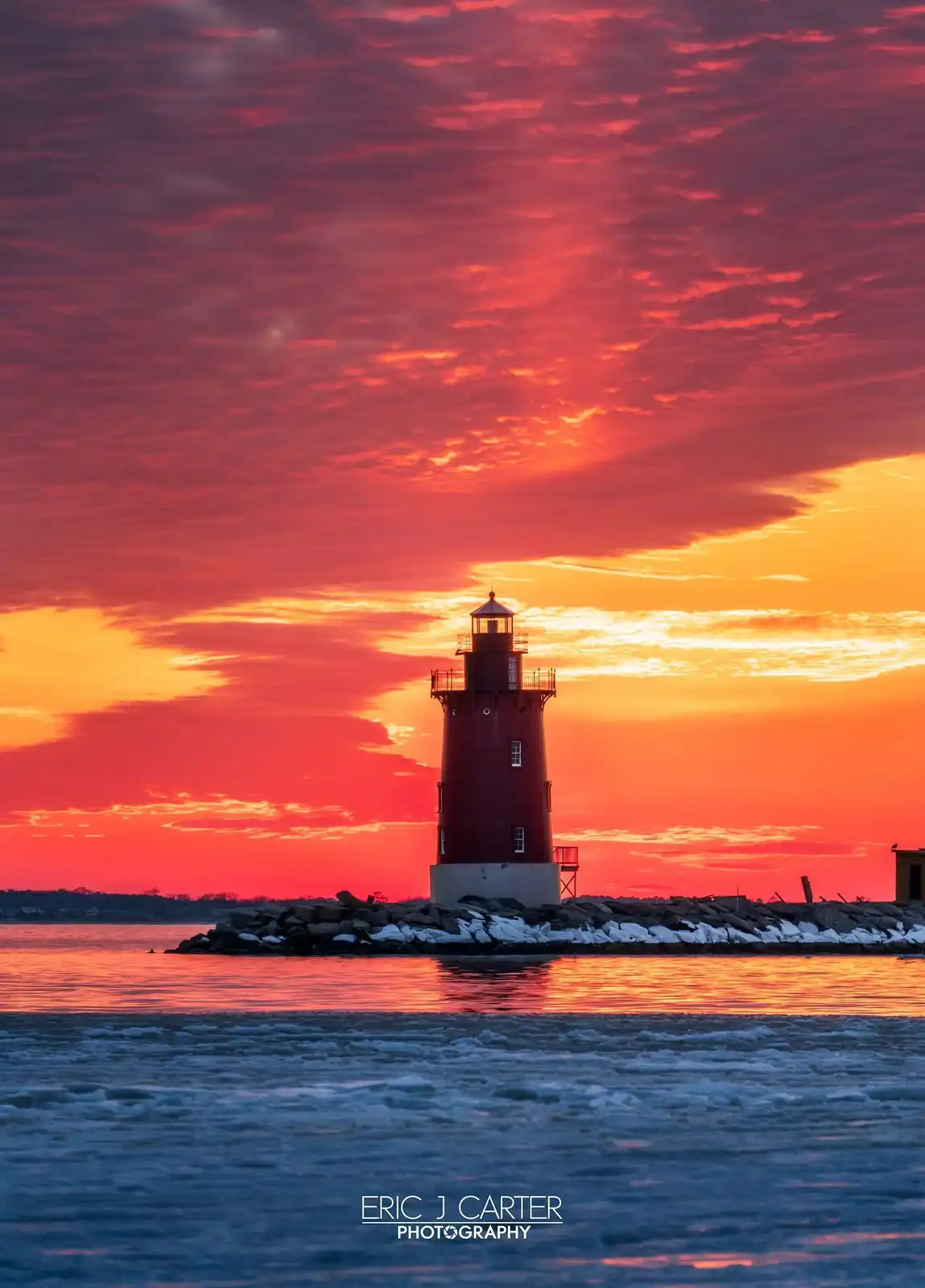 A red lighthouse stands on a rocky pier against a dramatic sunset sky filled with fiery red and orange clouds, with icy…