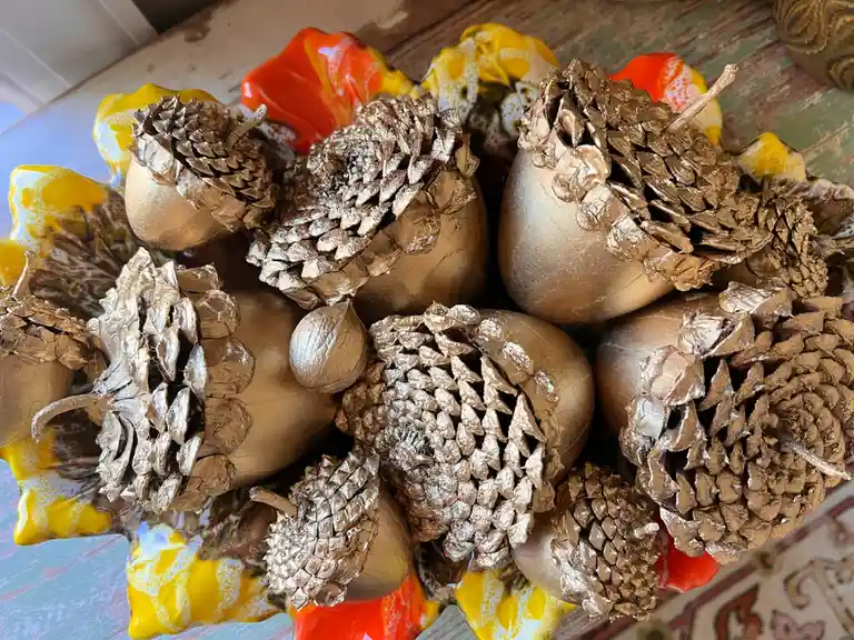 A close-up photo shows a decorative arrangement of pinecones and acorns, all coated in a shiny gold leaf finish, nestled…