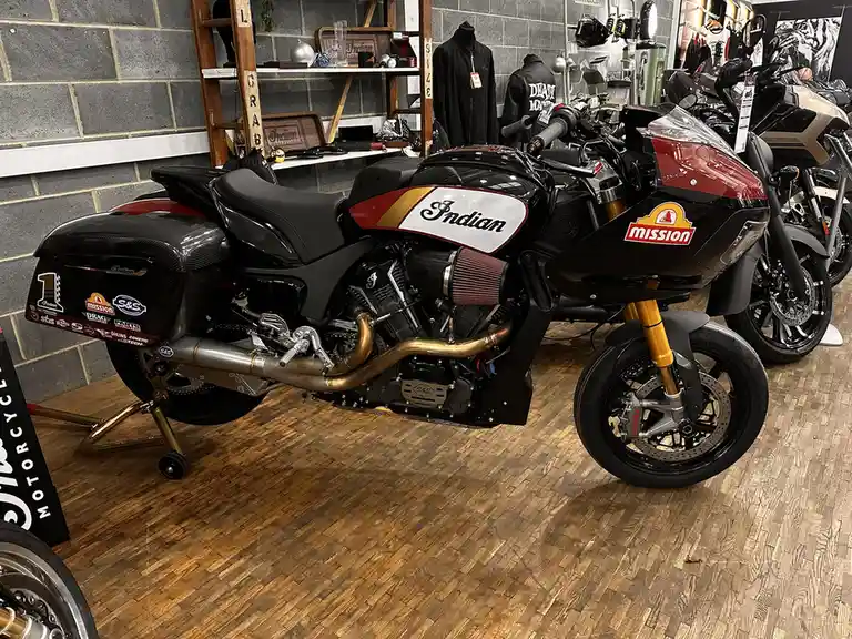 A black and red Indian motorcycle with gold accents and a 'mission' logo on the front fairing is displayed indoors on a…