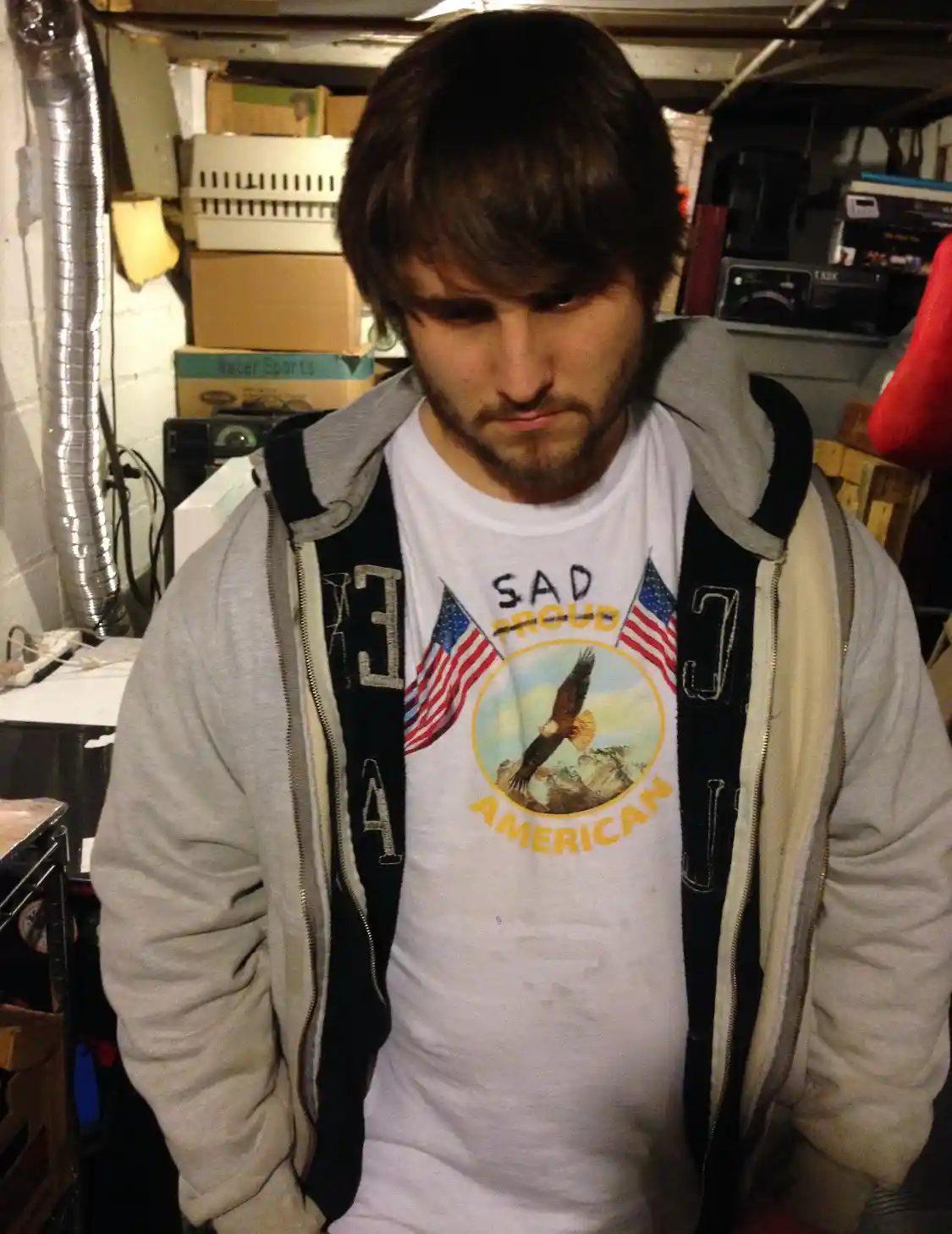 A man with a weary expression stands in a cluttered basement wearing a white t-shirt that reads 'SAD PROUD AMERICAN' with an…