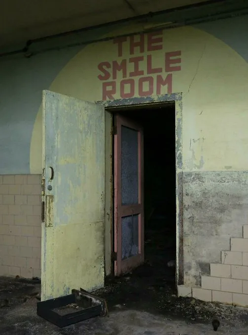 A dilapidated, abandoned doorway with peeling paint and debris on the floor, leading into darkness. Above the door, faded…