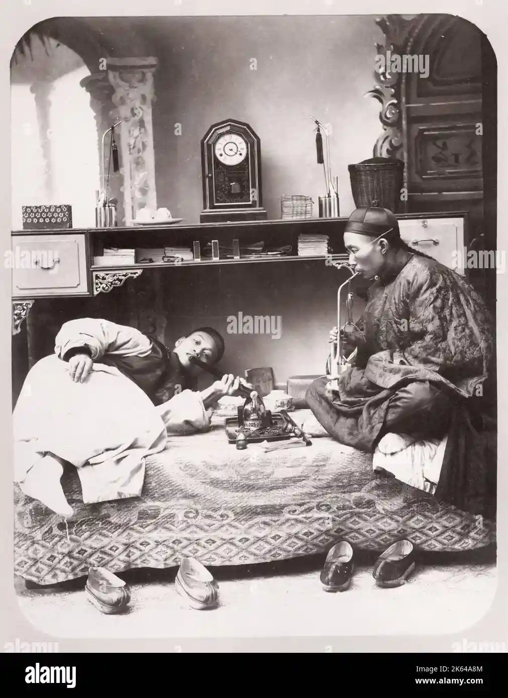 A vintage black-and-white photo shows two men in traditional Chinese attire sitting on a patterned rug. One man is propped…