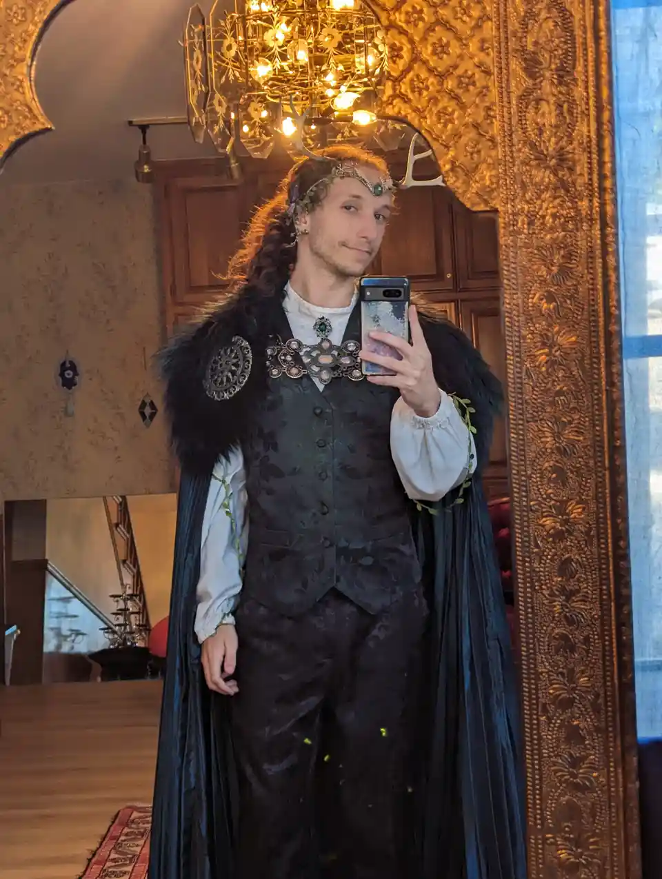 A person with long hair and antlers takes a mirror selfie in an ornate room, wearing a dark, elaborate costume with a…