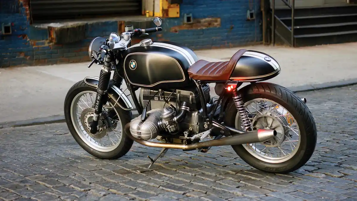 A custom-built black and silver BMW motorcycle with white racing stripes and a brown leather seat is parked on a cobblestone…