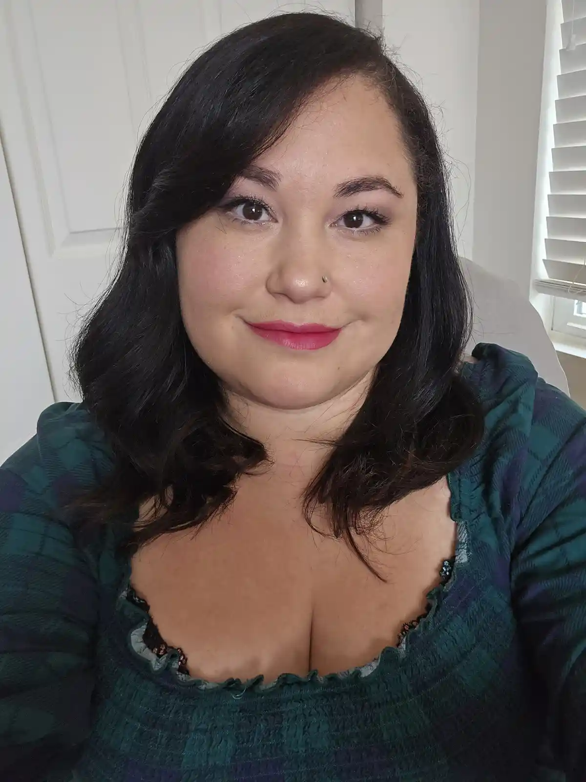 A woman with dark wavy hair and pink lipstick smiles at the camera while wearing a green plaid top with a ruffled neckline…
