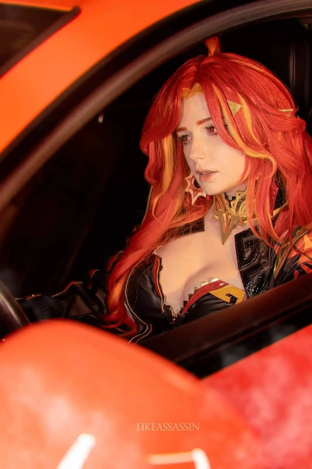 A person in a detailed cosplay of a character with long red and orange hair, wearing a black and gold outfit, sits inside an…