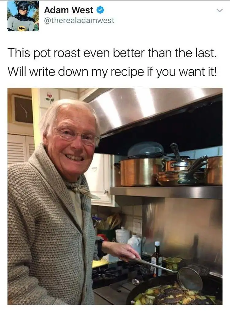 An older man with white hair and glasses, wearing a beige knit cardigan, smiles proudly while stirring a pot of food on a…