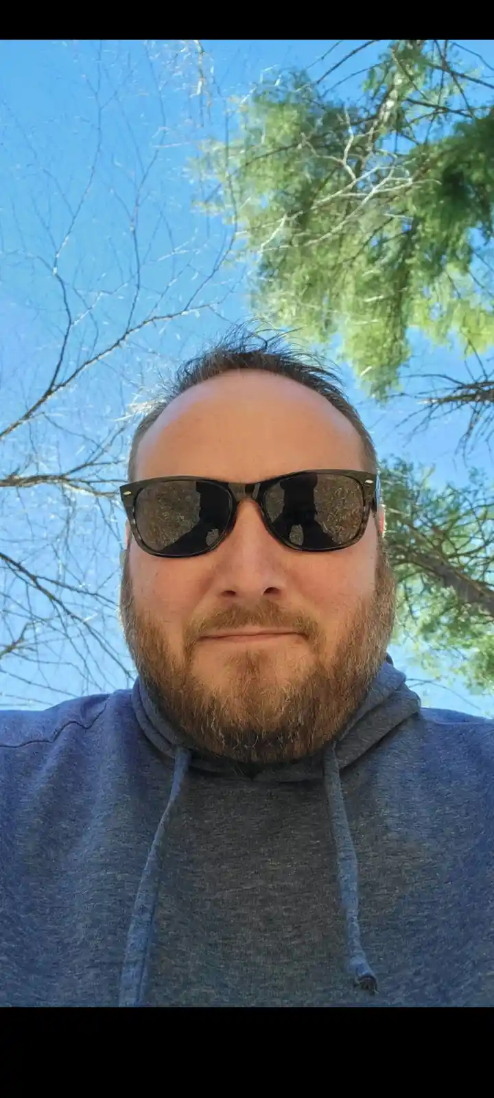 A man with a beard and sunglasses takes a selfie outdoors under a blue sky with trees, looking directly at the camera with a…