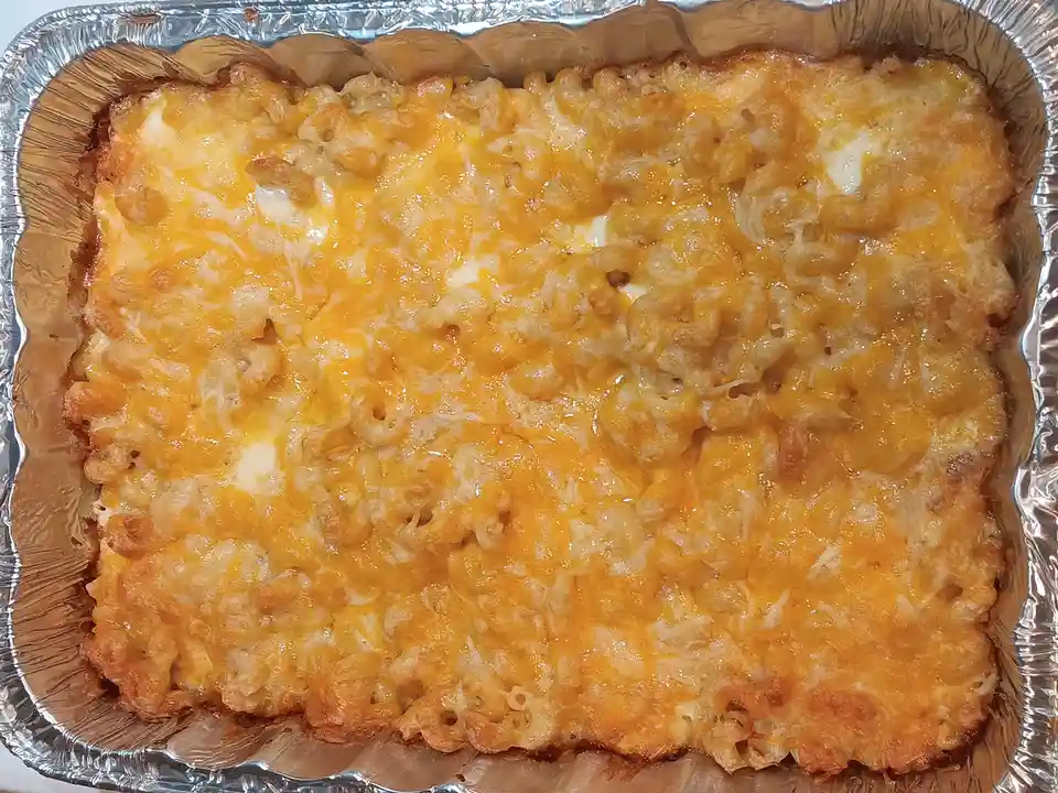 A close-up photo of a baked macaroni and cheese casserole in a rectangular aluminum foil baking pan, with a golden-brown…