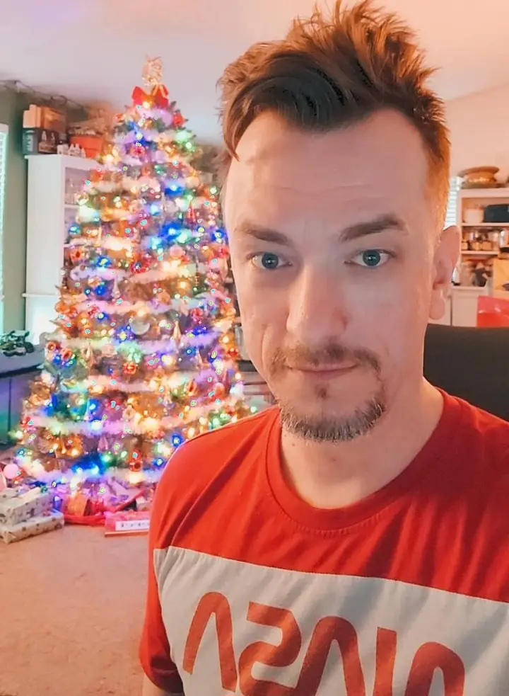 A man with spiky hair and a goatee stares directly at the camera with a deadpan expression, wearing a red t-shirt with…
