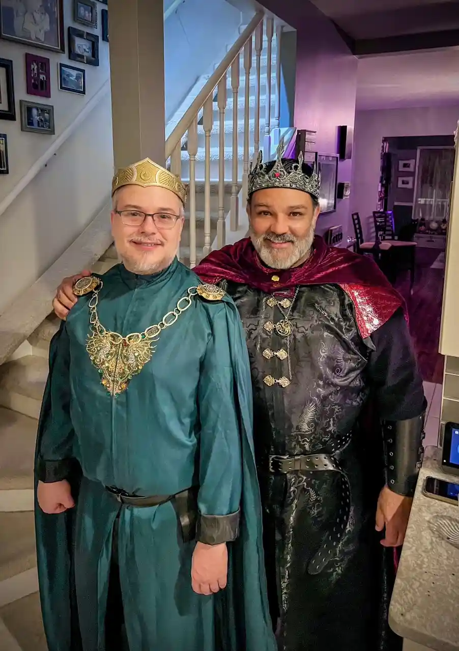 Two men in elaborate fantasy costumes pose indoors near a staircase; one wears a gold crown and teal robe with a jeweled…