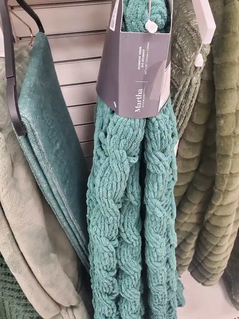 A teal, thickly braided decorative throw blanket by Martha Stewart hangs on a store rack, surrounded by other textured…