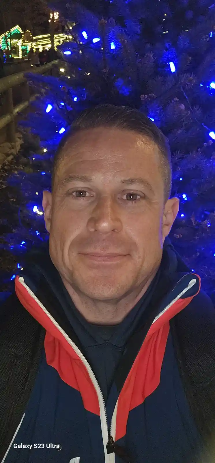 A man in a blue and red jacket smiles for a selfie at night, standing in front of a pine tree decorated with blue lights. In…