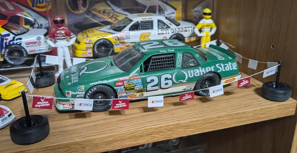 A green NASCAR model race car with number 26 and 'Quaker State' branding is displayed on a wooden shelf, surrounded by…