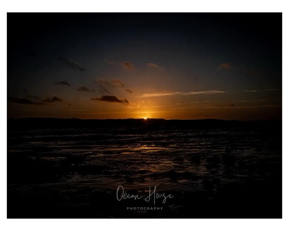 A dark, atmospheric photograph of a sunset over a body of water, with the sun low on the horizon casting orange light on…