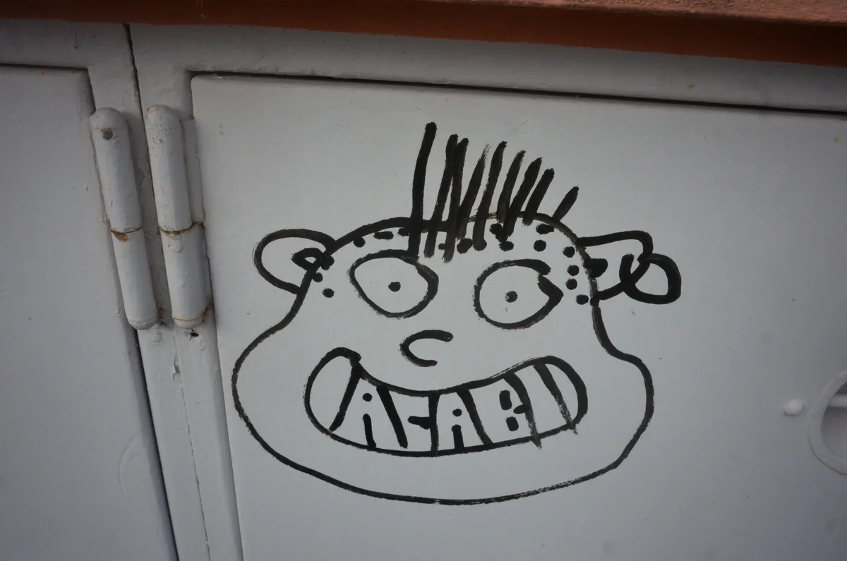 A crudely drawn cartoon face with spiky hair, wide eyes, and a toothy grin is painted in black on a white metal door. The…