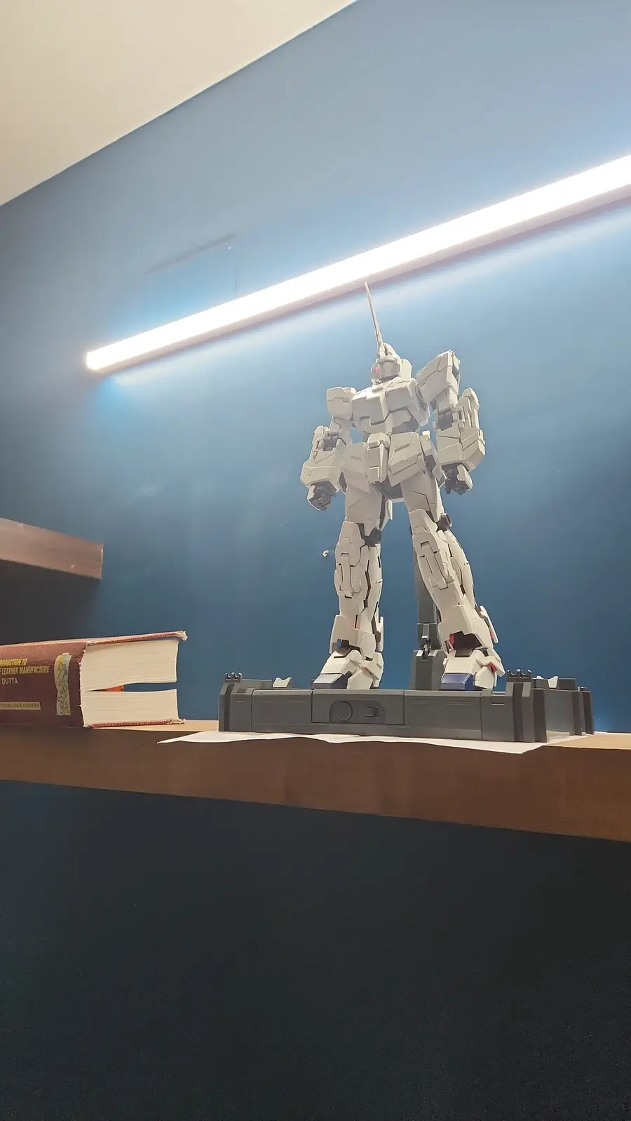 A white Gundam model, specifically the Unicorn Gundam, stands on a display stand next to a book with its pages torn out. The…