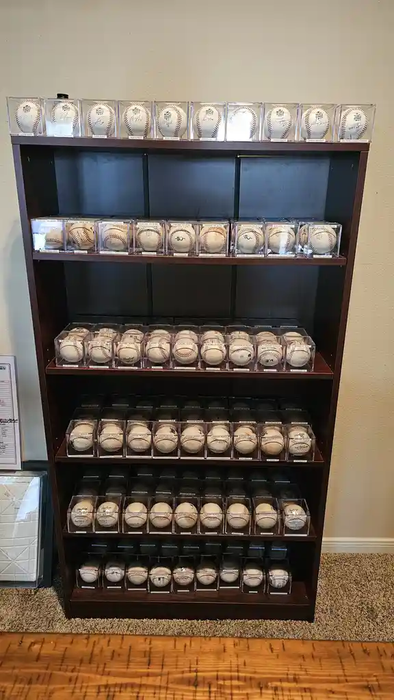 A tall wooden bookshelf is filled with dozens of baseballs, each individually housed in clear plastic display cases and…