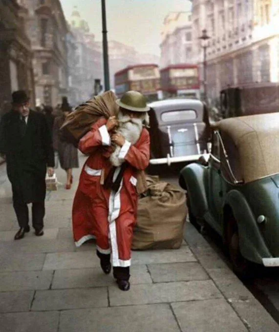 A man dressed as Santa Claus, wearing a red suit with white trim and a military-style helmet, is struggling to carry a large…