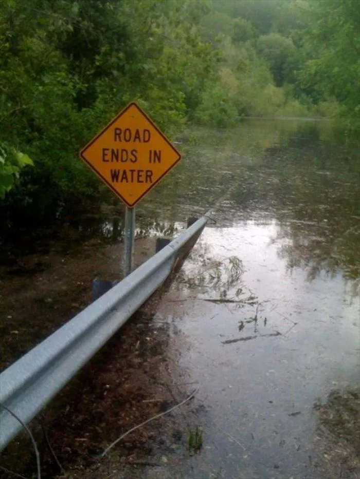 A yellow diamond-shaped road sign reading 'ROAD ENDS IN WATER' stands beside a flooded road, with water covering the…