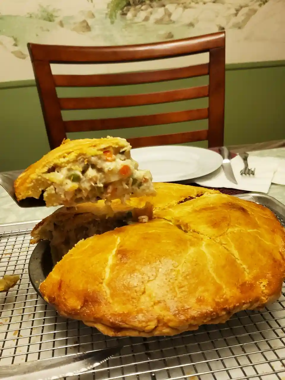 A large, golden-brown savory pie sits on a cooling rack, with a slice being lifted to reveal a creamy filling of meat and…