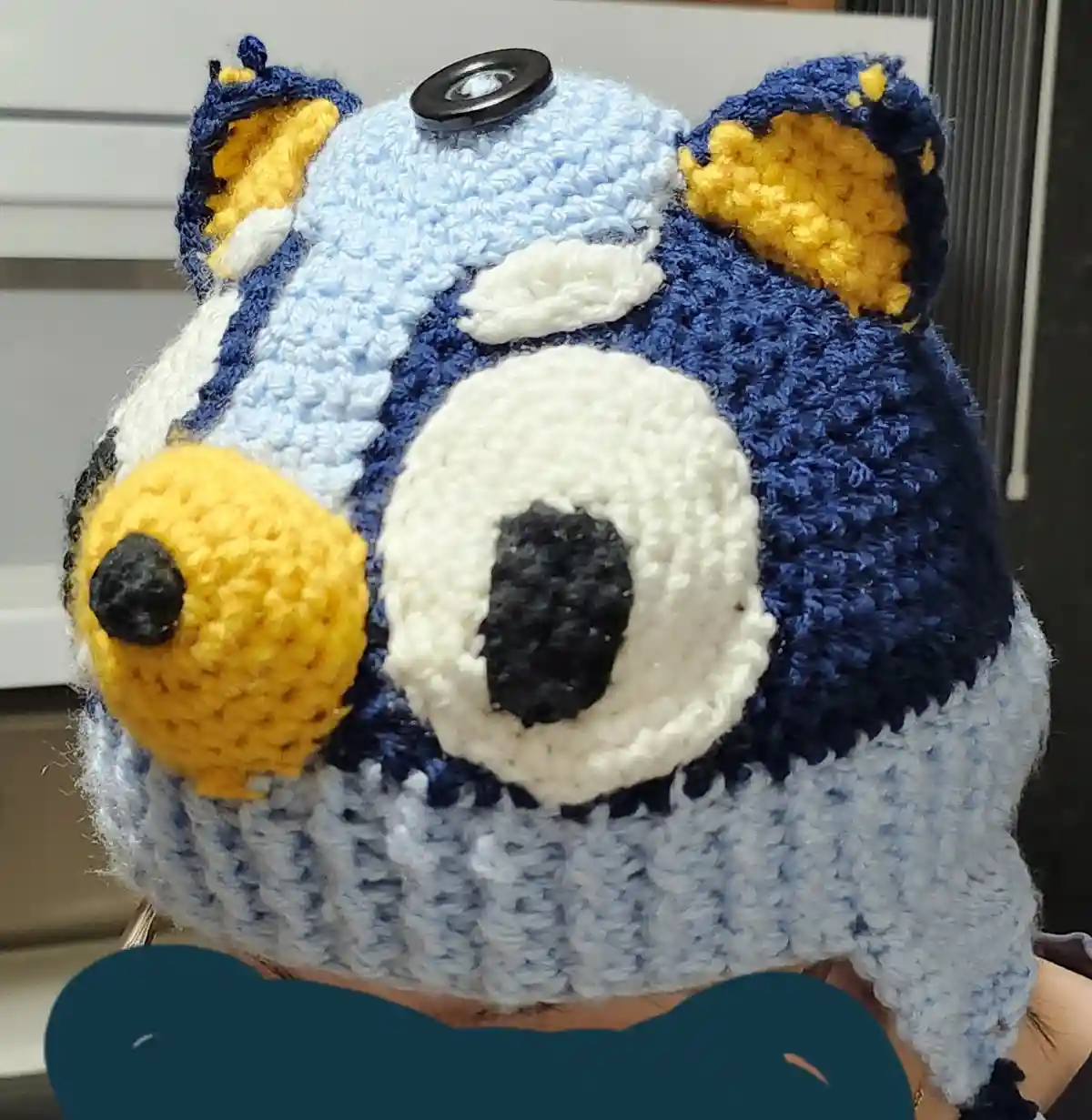 A close-up of a crocheted hat shaped like the cartoon dog Bluey, featuring blue, white, and yellow yarn, with a large white…