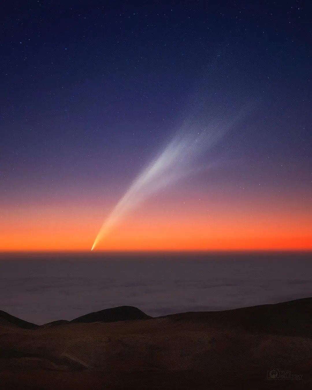 A stunning night sky photograph shows a bright, glowing comet with a long, curved tail arcing across a gradient sky…