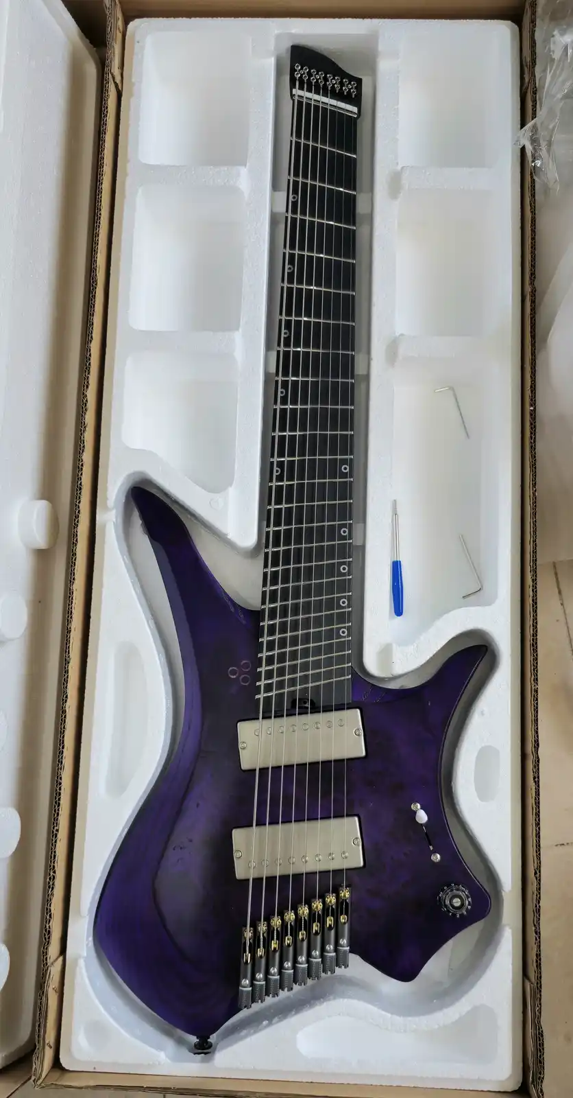 A purple electric guitar with seven strings is nestled in white foam packaging inside a cardboard box, accompanied by a…