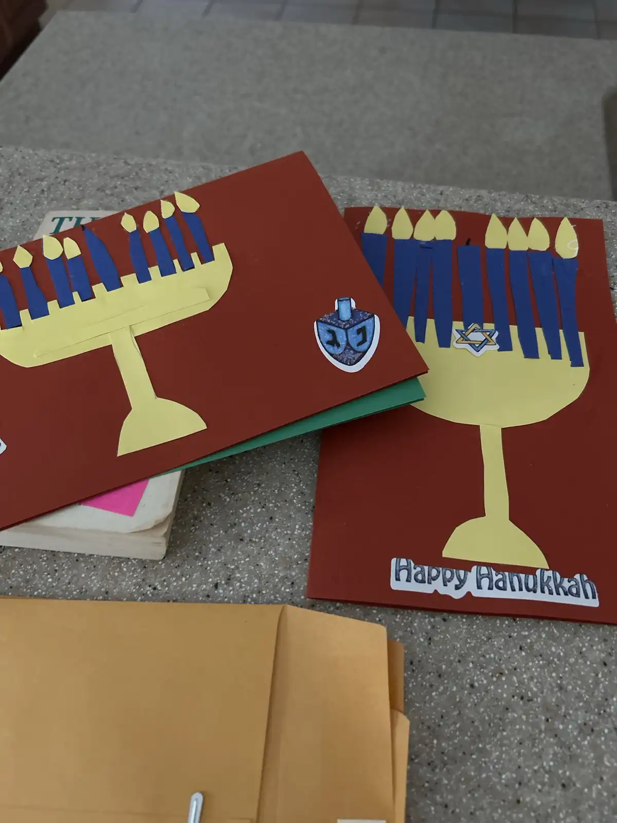 Two handmade Hanukkah greeting cards with paper cutouts of menorahs, blue candles, and holiday symbols like a dreidel and…