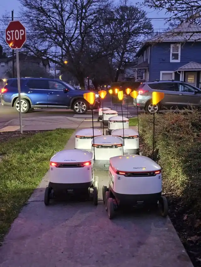 A row of white, self-driving delivery robots with glowing orange lights on their antennae are lined up on a sidewalk at…