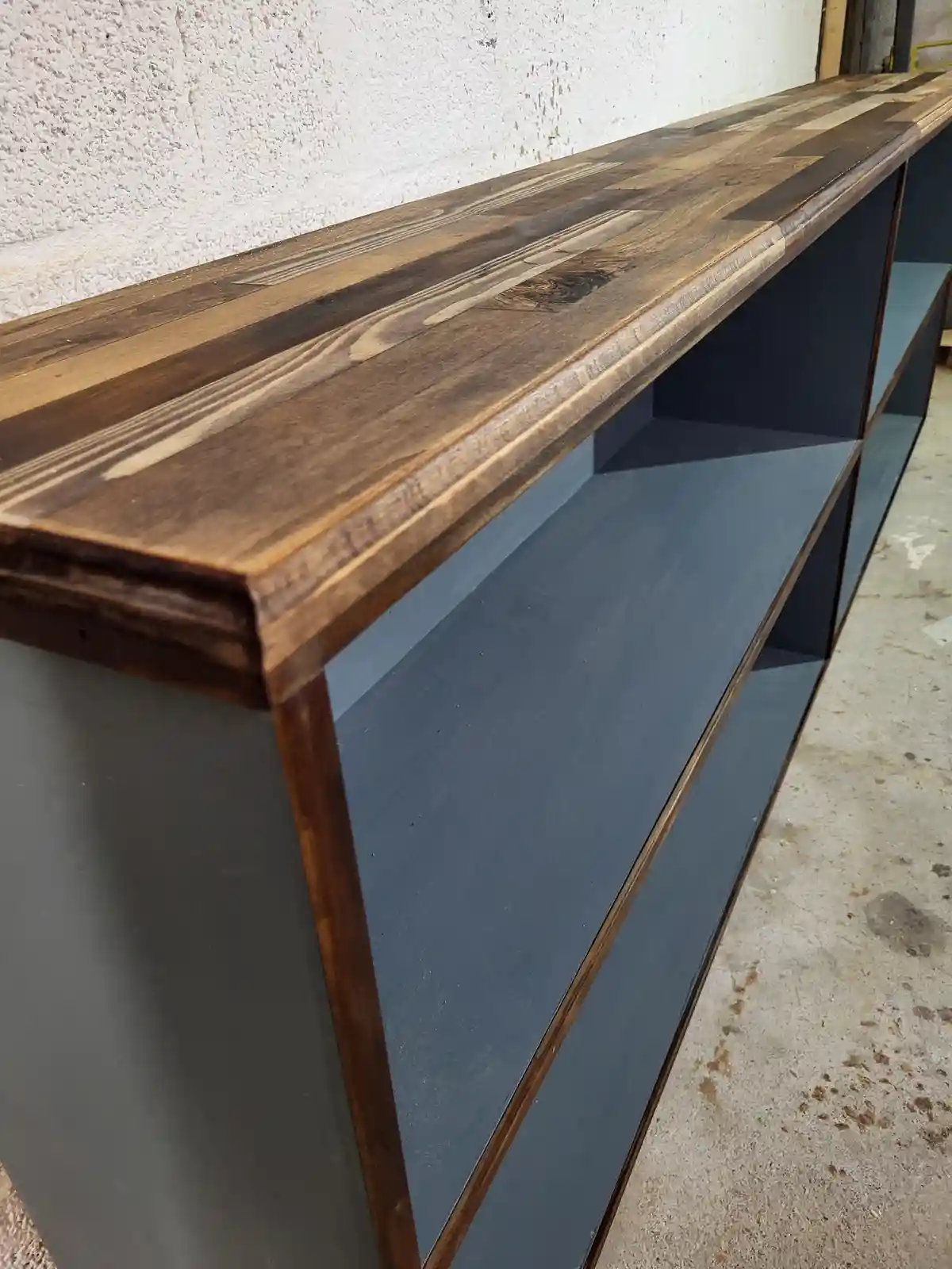A close-up photo of a modern shelf with a dark wood top and grey painted sides, set against a concrete wall. The overlaid…