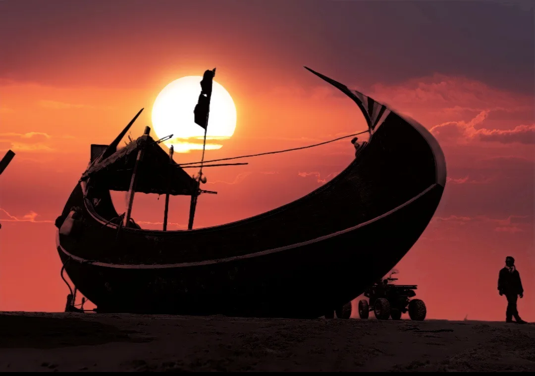 A silhouette of a large, curved boat with a flag and canopy sits on a sandy beach at sunset, with a small ATV parked beside…