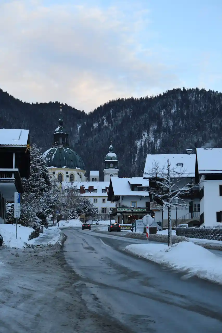 A snowy street in a European village with a large domed church and snow-covered buildings, cars driving on a partially…
